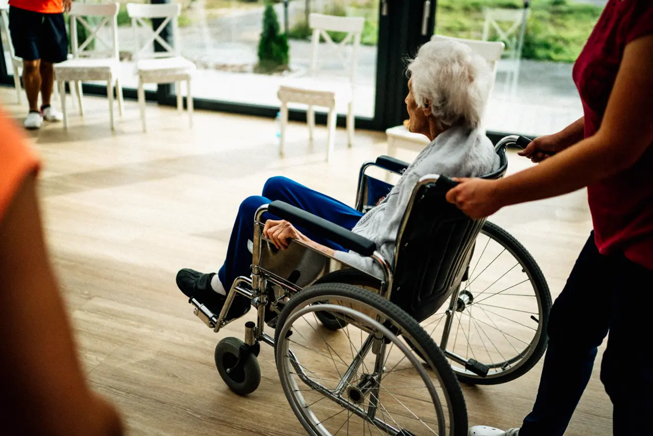 Seniorin im Rollstuhl an einem hellen Fenster, eine Pflegerin schiebt